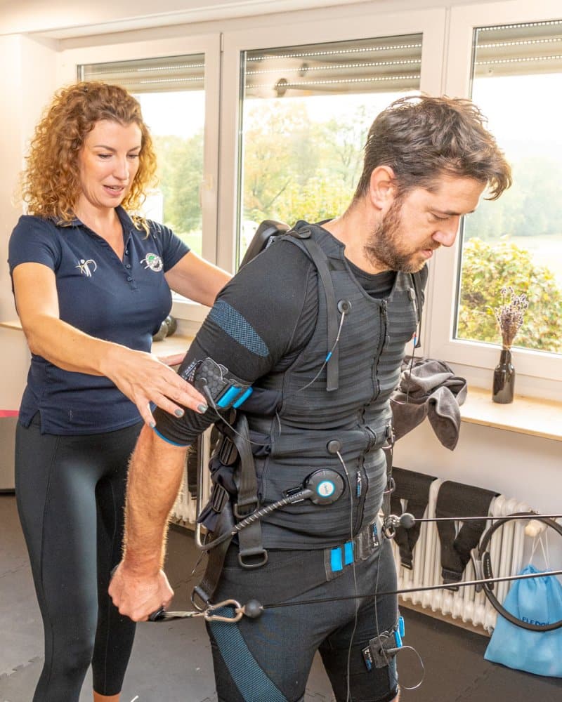 Man wearing a full-body rehabilitation/sensor suit with cables, assisted by a trainer in a bright gym.